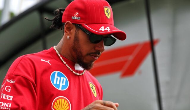 Ferrari driver wearing a red cap with number 44 and sunglasses, looking down with a beaded necklace on a red team shirt.