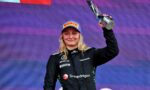 Female race car driver on a podium, holding up a trophy in a black racing suit with Snapdragon logo and a blue Pirelli cap.