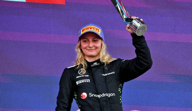 Female race car driver on a podium, holding up a trophy in a black racing suit with Snapdragon logo and a blue Pirelli cap.