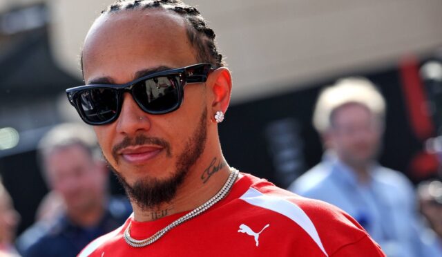 Man with braided hair wearing black sunglasses, a red Puma jersey, diamond necklace, and a necklace; outdoor portrait with tattoos visible on neck.",