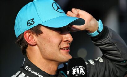 Close-up of a driver in a turquoise cap with a Mercedes logo, speaking into a FIA microphone, number 63 on the side of the cap.