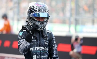 Formula 1 driver in Petronas racing suit and helmet raises a fist on the track with blurred grandstands behind him.