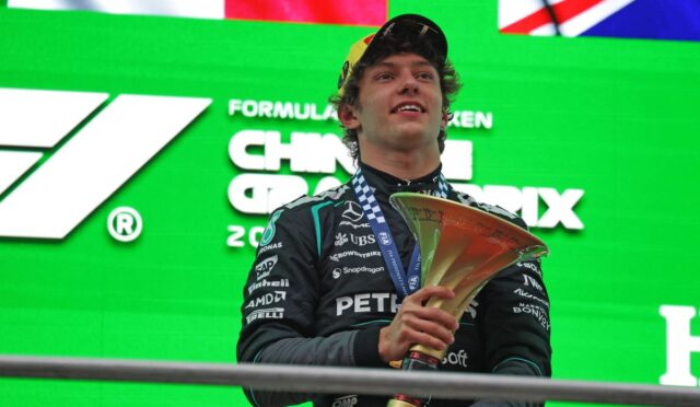 Young race car driver on a podium, holding a large gold trophy with a blue-and-white medal, in a dark racing suit against a bright green backdrop with sponsor logos.