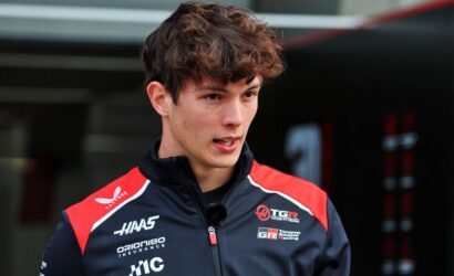 Portrait of a young male racing driver wearing a navy/red team jacket with sponsor logos, a small microphone clipped to his zipper, speaking or being interviewed in a pit area.