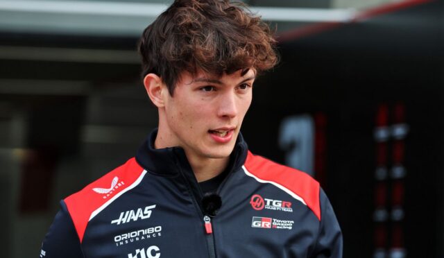 Portrait of a young male racing driver wearing a navy/red team jacket with sponsor logos, a small microphone clipped to his zipper, speaking or being interviewed in a pit area.