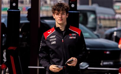 Young man in a black and red racing jacket holding race cards in a pit lane area.