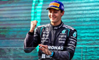 Mercedes race driver in a Petronas suit on a podium, smiling and raising a clenched fist in celebration.