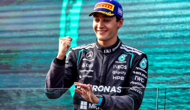 Mercedes race driver in a Petronas suit on a podium, smiling and raising a clenched fist in celebration.