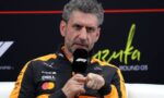 Man with gray curly hair in an orange-black racing jacket speaks into a microphone during an interview.