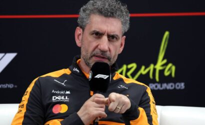 Man with gray curly hair in an orange-black racing jacket speaks into a microphone during an interview.