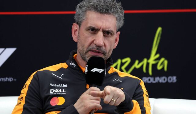 Man with gray curly hair in an orange-black racing jacket speaks into a microphone during an interview.