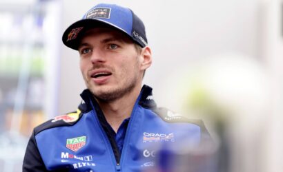 Male race car driver in a blue Oracle Red Bull Racing jacket and cap, speaking during an interview in a garage area.