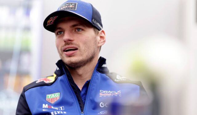 Male race car driver in a blue Oracle Red Bull Racing jacket and cap, speaking during an interview in a garage area.