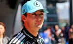 Portrait of a young race car driver wearing a turquoise Mercedes cap (with star logo) and a black TeamViewer racing suit, looking ahead outdoors.