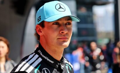 Portrait of a young race car driver wearing a turquoise Mercedes cap (with star logo) and a black TeamViewer racing suit, looking ahead outdoors.