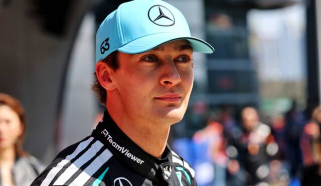 Portrait of a young race car driver wearing a turquoise Mercedes cap (with star logo) and a black TeamViewer racing suit, looking ahead outdoors.