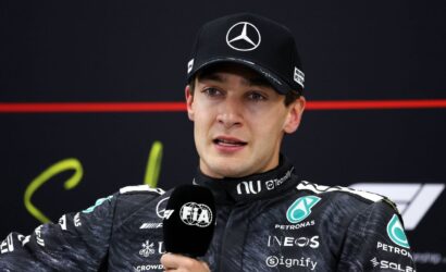 Male race car driver in a black Mercedes cap giving an interview, holding a FIA microphone with team logos visible on his racing suit behind him on a dark backdrop.