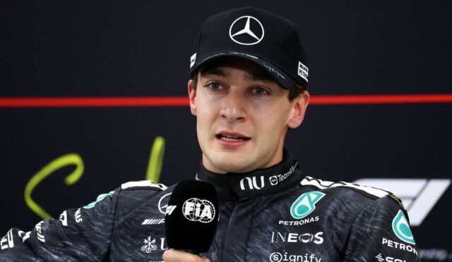 Male race car driver in a black Mercedes cap giving an interview, holding a FIA microphone with team logos visible on his racing suit behind him on a dark backdrop.