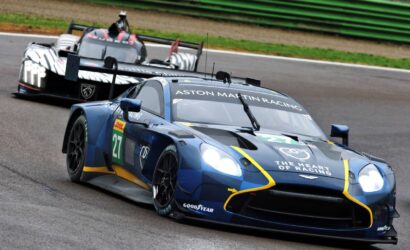 Blue Aston Martin Racing car #27 on a race track with a zebra-striped prototype close behind.