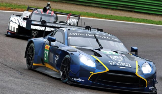 Blue Aston Martin Racing car #27 on a race track with a zebra-striped prototype close behind.