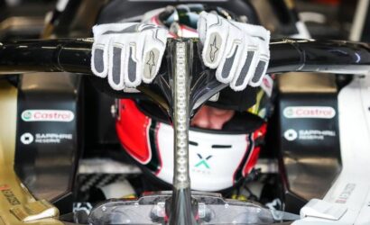 Race car driver in helmet with white racing gloves draped over the cockpit roll hoop of an open-wheel race car.