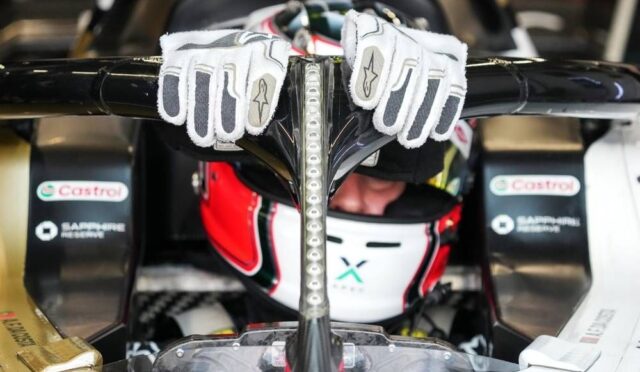 Race car driver in helmet with white racing gloves draped over the cockpit roll hoop of an open-wheel race car.