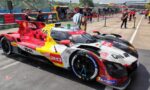 Colorful race car with red, yellow, and white livery parked on pit lane; visible sponsor logos and number 20 on the side. In the background, crew and garages along the track.