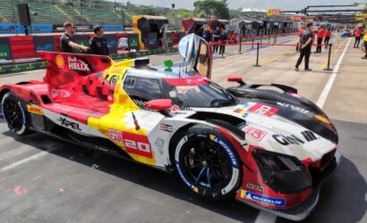 Colorful race car with red, yellow, and white livery parked on pit lane; visible sponsor logos and number 20 on the side. In the background, crew and garages along the track.