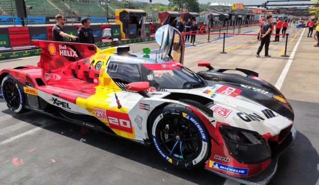 Colorful race car with red, yellow, and white livery parked on pit lane; visible sponsor logos and number 20 on the side. In the background, crew and garages along the track.