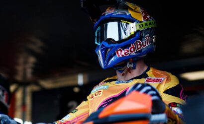 Motocross rider in a yellow jersey and blue Red Bull helmet with mirrored goggles sits on an orange bike in a dim pit area.