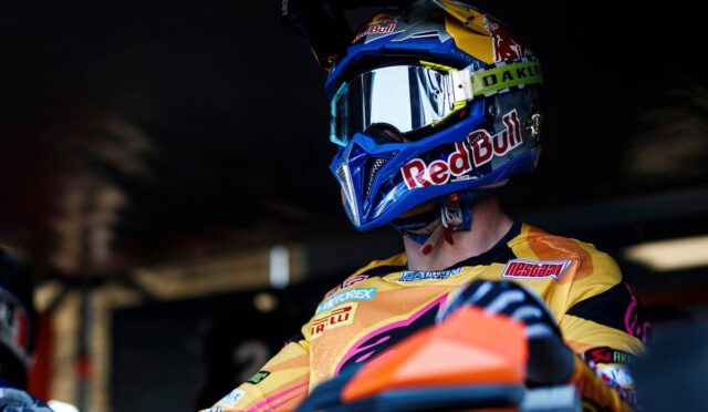 Motocross rider in a yellow jersey and blue Red Bull helmet with mirrored goggles sits on an orange bike in a dim pit area.