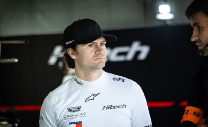 Race car driver in a white suit and cap stands in a garage, listening with a focused expression next to a teammate wearing a headset.