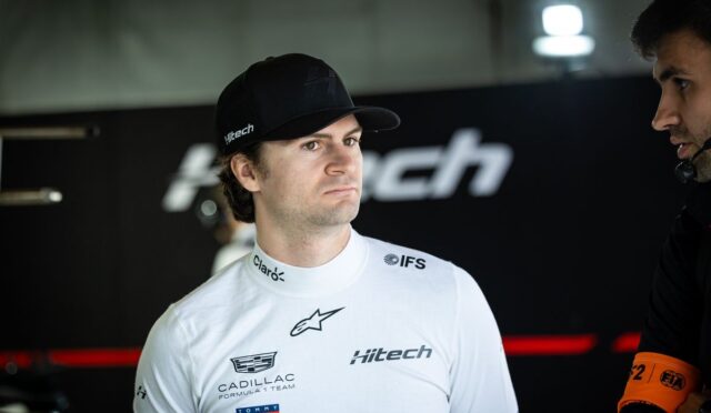 Race car driver in a white suit and cap stands in a garage, listening with a focused expression next to a teammate wearing a headset.