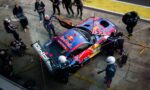 Top-down view of a Red Bull racing car in the pit lane surrounded by crew members during a pit stop; crew change tires and refuel as photographers and cameras surround the scene