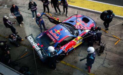 Top-down view of a Red Bull racing car in the pit lane surrounded by crew members during a pit stop; crew change tires and refuel as photographers and cameras surround the scene