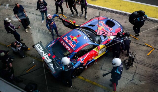 Top-down view of a Red Bull racing car in the pit lane surrounded by crew members during a pit stop; crew change tires and refuel as photographers and cameras surround the scene