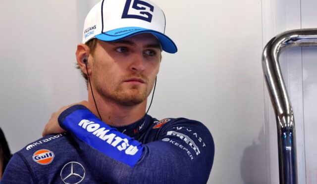 Male race car driver in a blue Williams racing suit and white cap with a blue bill, wearing earbuds and resting his arm on his shoulder in a garage.