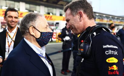 Two men greet each other in a racing pit area: one wearing a suit and a colorful patterned face mask, the other in a dark racing uniform with a headset and sponsor logos (Red Bull, Citrix).