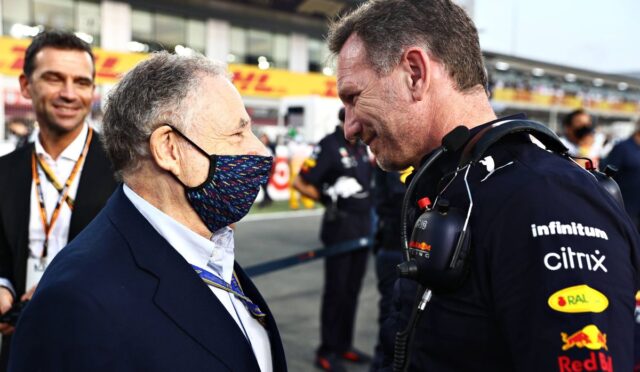 Two men greet each other in a racing pit area: one wearing a suit and a colorful patterned face mask, the other in a dark racing uniform with a headset and sponsor logos (Red Bull, Citrix).