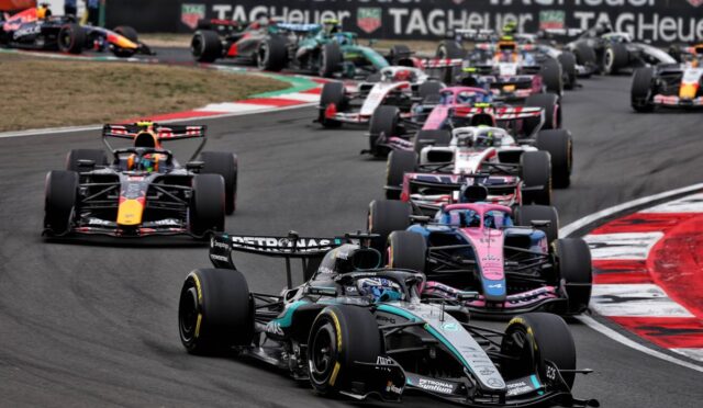 Multiple Formula 1 cars race closely around a curve; Mercedes leads with TAG Heuer banners in the background.