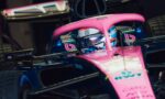 Pink and blue Formula 1 car with driver in cockpit peering over the visor inside a garage.