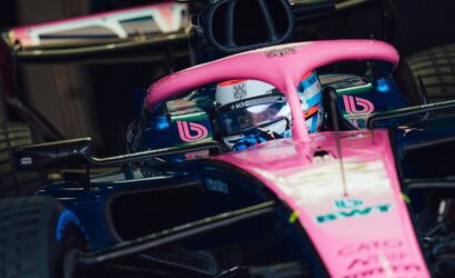 Pink and blue Formula 1 car with driver in cockpit peering over the visor inside a garage.