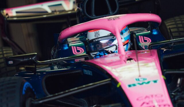 Pink and blue Formula 1 car with driver in cockpit peering over the visor inside a garage.