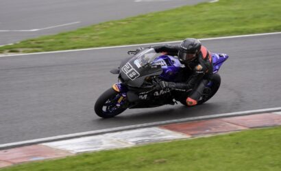 Motorcycle racer in a black suit and helmet leans into a left-hand turn on a blue/purple sport bike on a race track with green grass nearby.