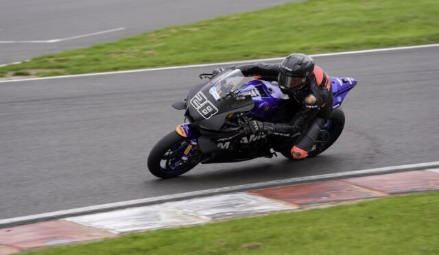 Motorcycle racer in a black suit and helmet leans into a left-hand turn on a blue/purple sport bike on a race track with green grass nearby.