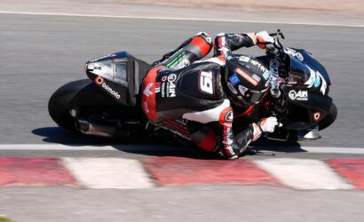 Moto racer in red–black leathers leans into a tight turn on a black race bike with sponsor logos, crossing red‑white track curbs.