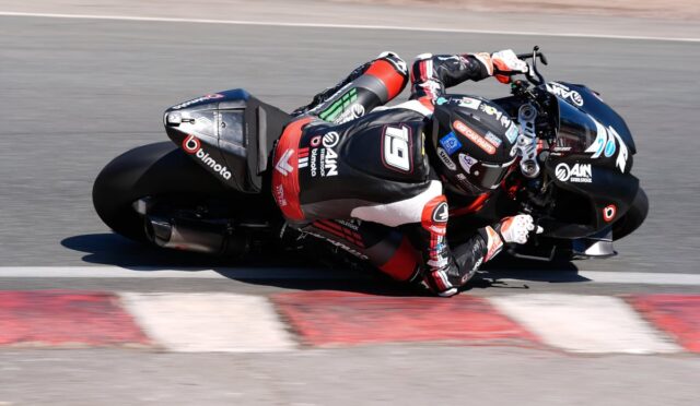 Moto racer in red–black leathers leans into a tight turn on a black race bike with sponsor logos, crossing red‑white track curbs.