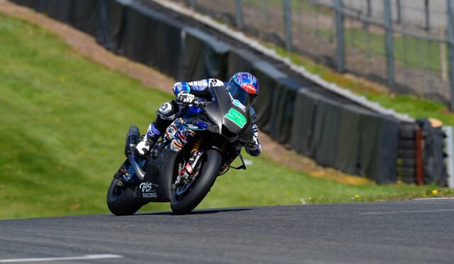 Motorcycle racer in blue gear leaning into a turn on a race track, black bike with green '52' number plate.