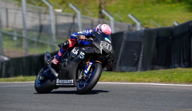 Motorcycle racer in a pink helmet leans into a turn on a race track, blue and black bike numbered 28 in view