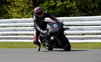 Motorcycle racer in black and red leathers leaning into a turn on a racing bike with the number 43 on the front fairing, on a track with a metal guardrail in the background.